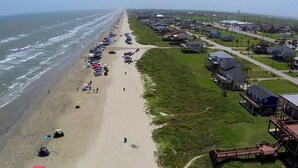 Une plage bordée de rangées de parasols et où sont garées des voitures le long du rivage. Des maisons et une route se trouvent à proximité de la plage.