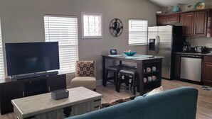 Un salon avec un téléviseur, un coin repas avec une table et des chaises, une cuisine équipée et une grande fenêtre avec des stores.