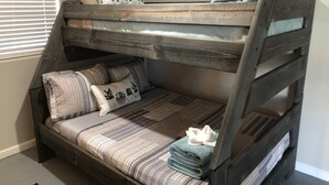Un lit superposé avec un matelas et des oreillers à rayures, une petite fenêtre avec des stores et un mur orné d’un tableau floral.