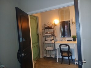 A room with a wooden door, a metal shelf with folded towels, a chair, a small table, and a mirror with a light.