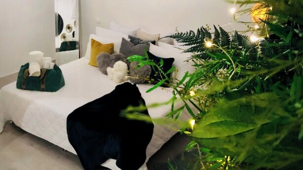 Een slaapkamer met een bed, kussens, een spiegel en een kerstboom met lichtjes.