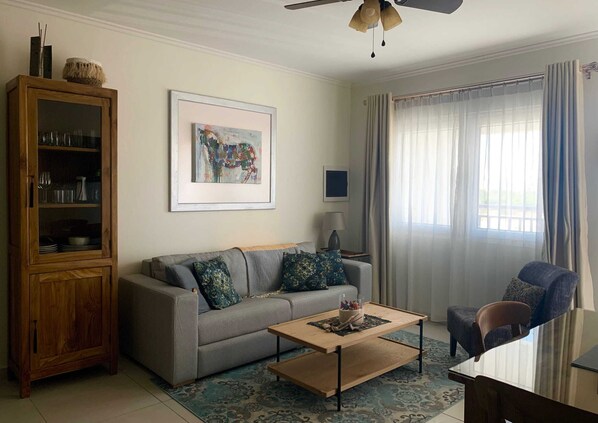 A living room with a grey sofa, a wooden coffee table, a cabinet, a chair, and a large window with curtains. - Gladiola Arena Condo at Eagle Beach! (Noord)