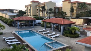 A pool area with lounge chairs and a covered seating area, surrounded by palm trees and buildings. - Gladiola Arena Condo at Eagle Beach! (Noord)