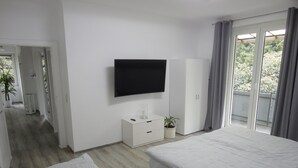 Ein modernes Hotelzimmer mit zwei Betten, einem Fernseher, einer kleinen Kommode, einer Topfpflanze und einem Balkon mit Vorhängen.