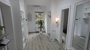 Ein Flur mit Holzboden, weißen Wänden und einem Blumentopf. Auf beiden Seiten befinden sich Türen sowie ein kleiner Tisch mit einer Lampe.