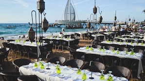 An outdoor dining area with tables set for guests, overlooking the sea and a bridge. - La Palapa beachfront fully renovated condo!  In the heart of the Romantic Zone. (Puerto Vallarta)