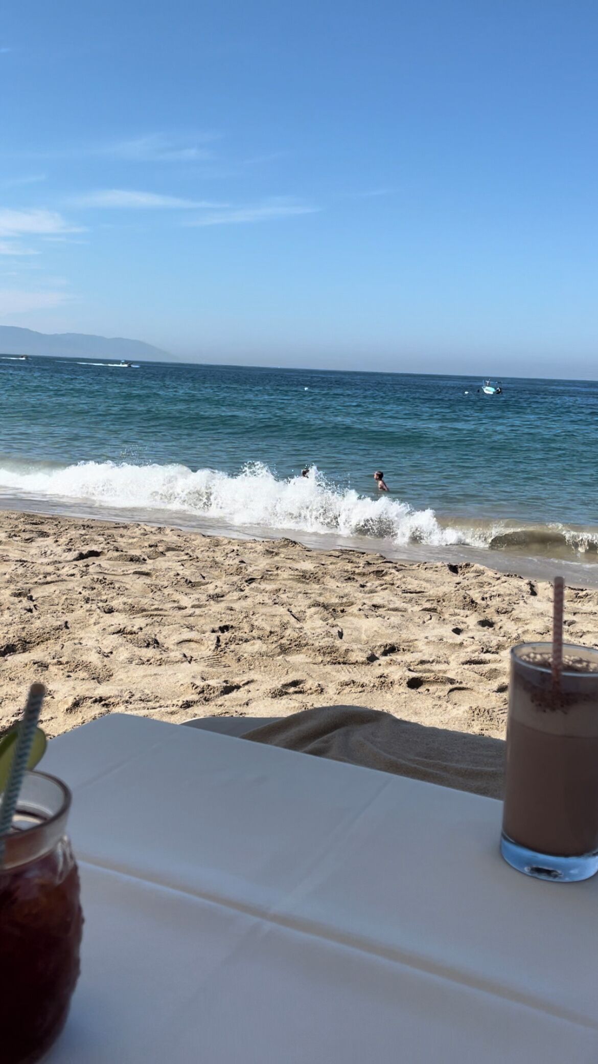 Une vue sur la plage avec une table et des boissons, les vagues qui se brisent sur le rivage et un bateau au loin.