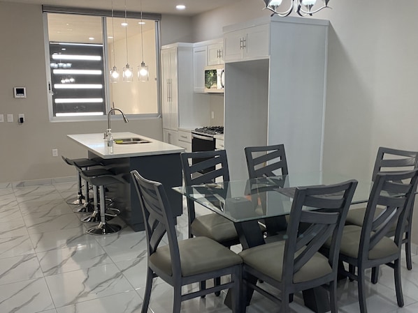 A modern kitchen with a central island, a dining area with six chairs, and a large window with pendant lighting.