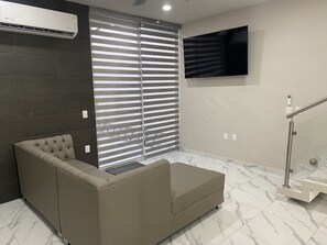 A modern living room with a beige sofa, a large window with blinds, a mounted TV, and a staircase with a glass railing.