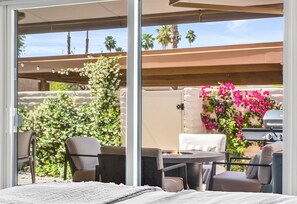 An outdoor patio with a grill, seating, and a view of a garden with flowers and palm trees.