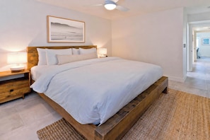 Un dormitorio con estructura de cama de madera, ropa de cama blanca, dos lámparas de noche, un ventilador de techo y un cuadro en la pared.