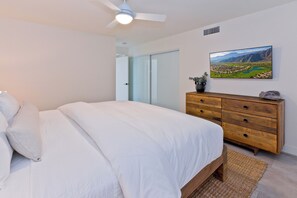 A bedroom with a bed, a wooden dresser, a ceiling fan, a wall-mounted TV, and a door leading to another room.