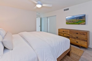 Une chambre à coucher avec un lit, une commode en bois, un ventilateur de plafond et un tableau accroché au mur.