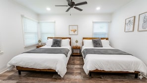 Une chambre avec deux lits, des têtes de lit en bois, du linge de lit blanc, un ventilateur de plafond et des œuvres d’art encadrées accrochées aux murs.