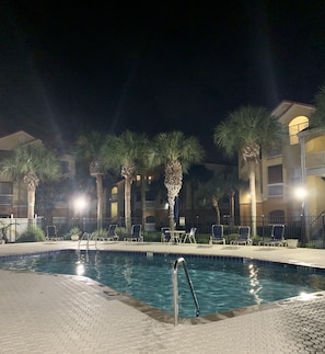 A nighttime swimming pool area with lounge chairs and palm trees, surrounded by multi-story buildings with balconies.