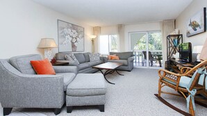 A living room with grey sofas, a wooden coffee table, and a wicker rocking chair. There is a TV on a stand, a lamp, and a painting on the wall.