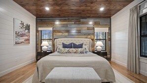 A bedroom with a large bed, wooden ceiling, two bedside tables with lamps, a window with curtains, and a painting on the wall. - 3 Masters, w/Bunk Room, Shuffle Board, Arcades, Hot Tub-Great For Families (Broken Bow)