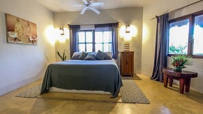 Ein Schlafzimmer mit einem Bett, einem Deckenventilator, einem Fenster mit Vorhängen, einem Nachttisch und einer Wandlampe.