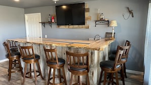 A bar area with a wooden bar top, six wooden bar stools, a flat-screen TV mounted above the bar, and a small shelf with bottles and a lamp.