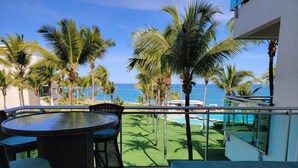 Ein Balkon mit rundem Tisch und Stühlen, Blick auf einen Pool und Palmen, mit Meerblick.