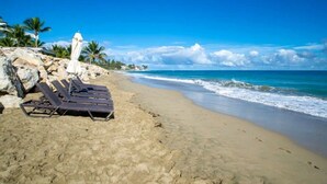 Ein Strand mit Liegestühlen und einem weißen Sonnenschirm, eine felsige Küstenlinie und Palmen im Hintergrund.