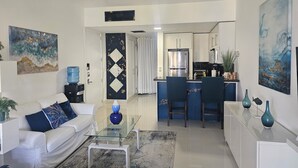 Ein modernes Wohnzimmer mit Glastisch, einer Couch und einer Küchenzeile inklusive Kühlschrank und Barhocker.
