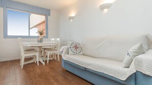Una habitación con sofá blanco, una mesa de comedor con sillas, una ventana con vistas y una lámpara fijada a la pared.