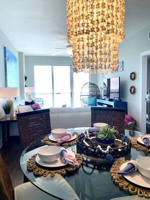 Uma área de refeições com mesa de vidro posta para quatro pessoas, rodeada por cadeiras de vime. Uma luminária decorativa pendurada está sobre a mesa.