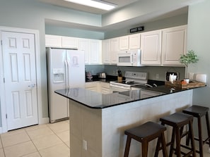 Uma cozinha moderna com armários brancos, uma ilha central com banquetas, geladeira, micro-ondas e fogão.