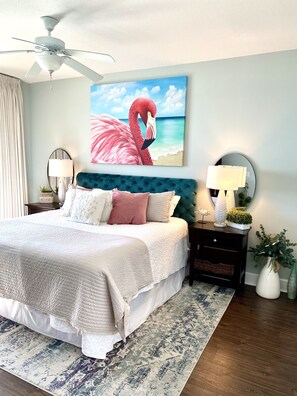A bedroom with a bed, a ceiling fan, a painting of a flamingo, a nightstand, a lamp, and a vase with plants. - Beachfront~Amazing View~Great Location~Beach Chair Service Included  (Panama City)