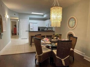 A dining area with a chandelier, wicker chairs, and a kitchen in the background.