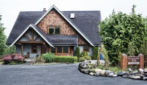Une maison en bois à deux étages, dotée d’un toit à forte pente, d’un porche à l’avant et d’une allée menant à un panneau en bois indiquant le nom et l’adresse de la propriété.