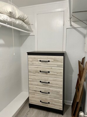 Una habitación blanca con un mueble de madera con cajones y tiradores negros, una puerta blanca y un estante blanco con almohadas.