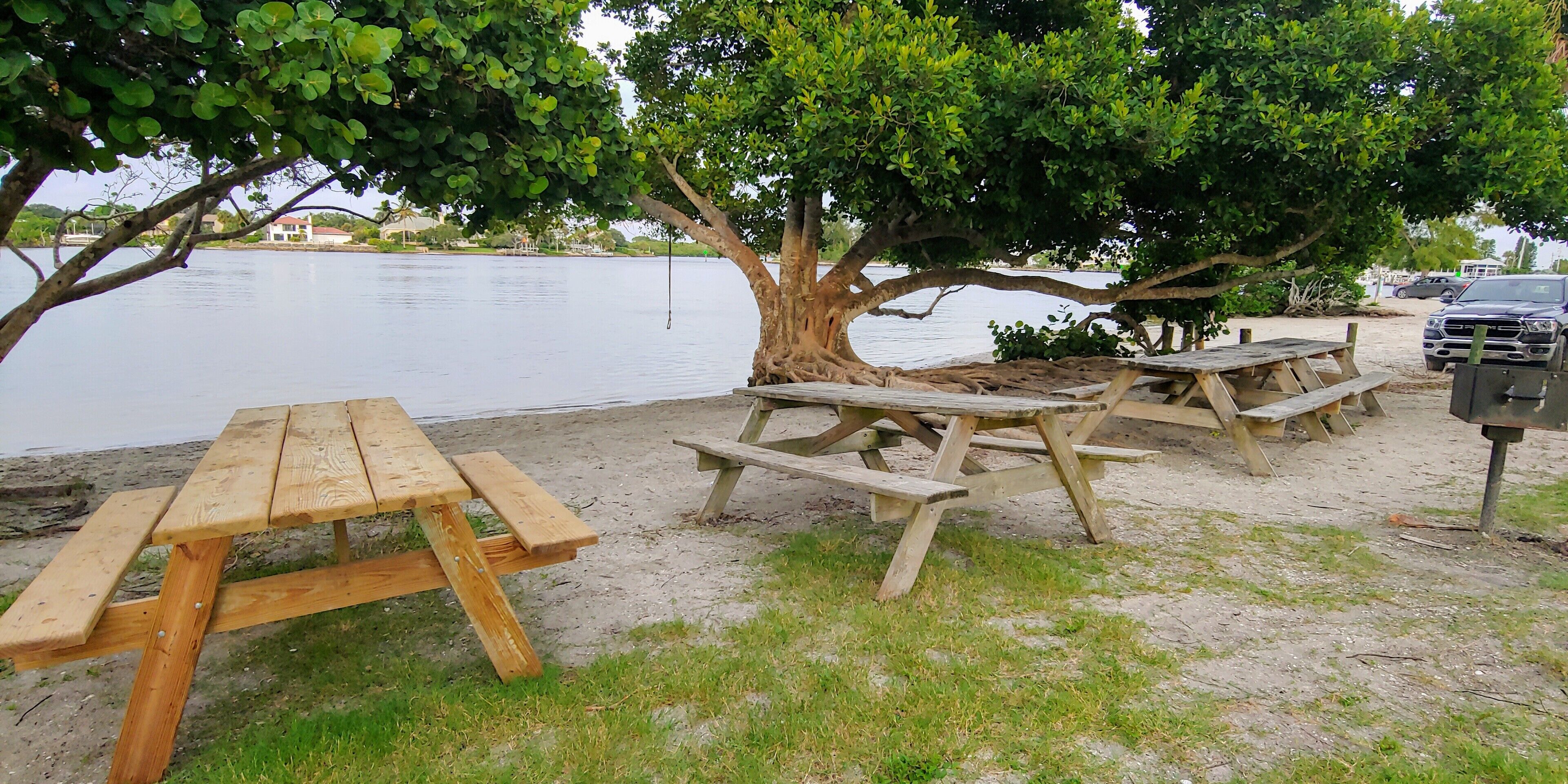 A lakeside picnic area with wooden benches and picnic tables, a parking area with a black SUV, and a large tree providing shade.