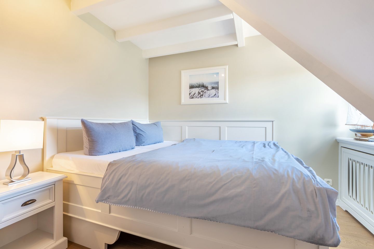 Een slaapkamer met een bed, nachtkastje en een ingelijst beeld aan de muur.