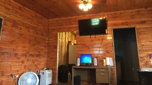 Ein Zimmer mit Holzvertäfelung, Deckenventilator, einem angebrachten Fernseher, einem Schreibtisch mit Computer und einem Standventilator.