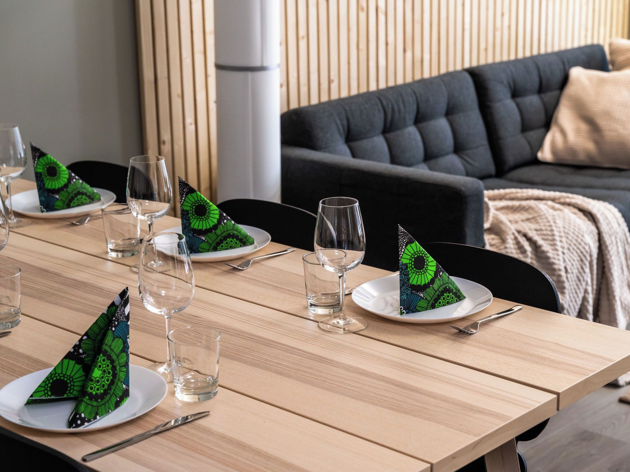 Una mesa de comedor dispuesta con platos, vasos y cubiertos, con un sofá y un muro de madera al fondo.