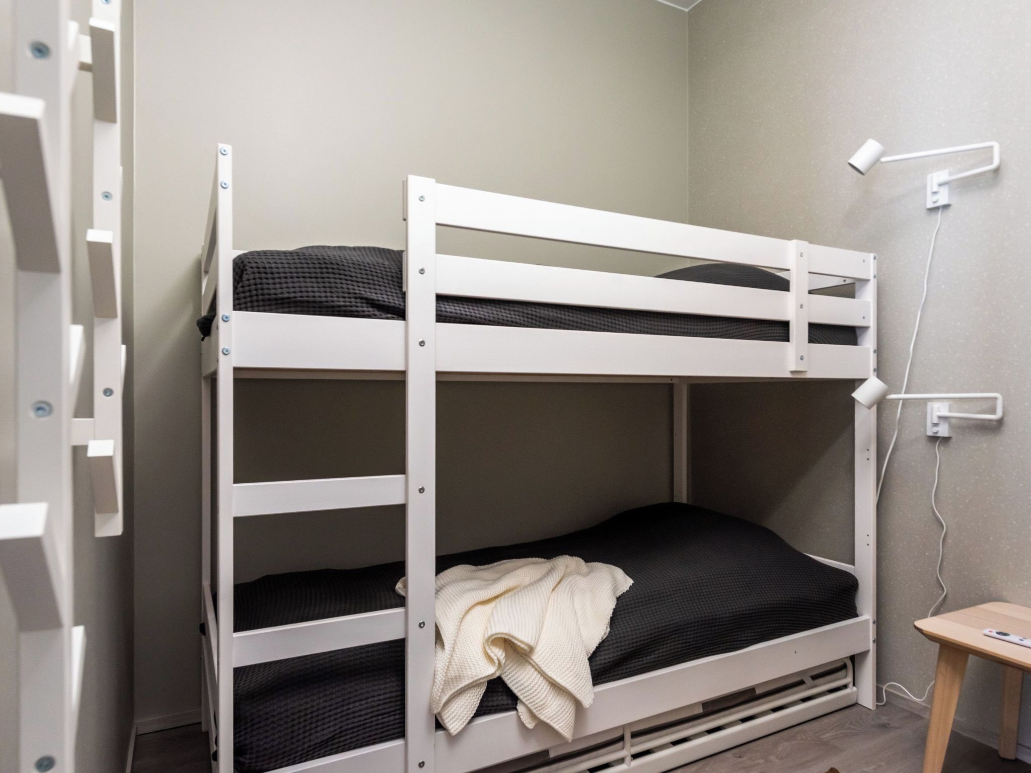Una cama litera con estructura blanca, una mesita pequeña y un aplique de pared.