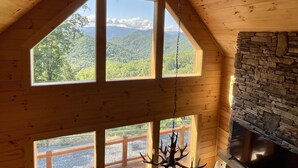 Ein Holzinnenraum mit Kamin aus Stein, großen Fenstern mit Blick auf die Berge und einer Kristallleuchter.