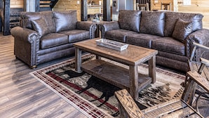 Ein geräumiges Wohnzimmer mit einer großen Holzbalkendecke, einem Kronleuchter, einem ledernen Ecksofa und einer Treppe, die in ein Obergeschoss führt.