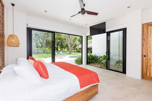 Un dormitorio con una cama, un ventilador de techo y amplias puertas correderas de cristal que dan a un jardín.
