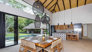 Un vaste espace de salle à manger avec une grande table en bois et des chaises en osier, une cuisine moderne équipée d’appareils électroménagers en acier inoxydable, et une vue sur un jardin luxuriant et une piscine, visible à travers de grandes portes coulissantes.
