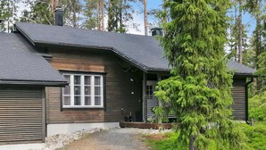 A modern log cabin with a gravel driveway and a large evergreen tree in front.