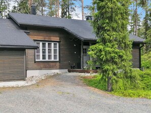 A two-story wooden cabin with a gravel driveway, surrounded by trees.