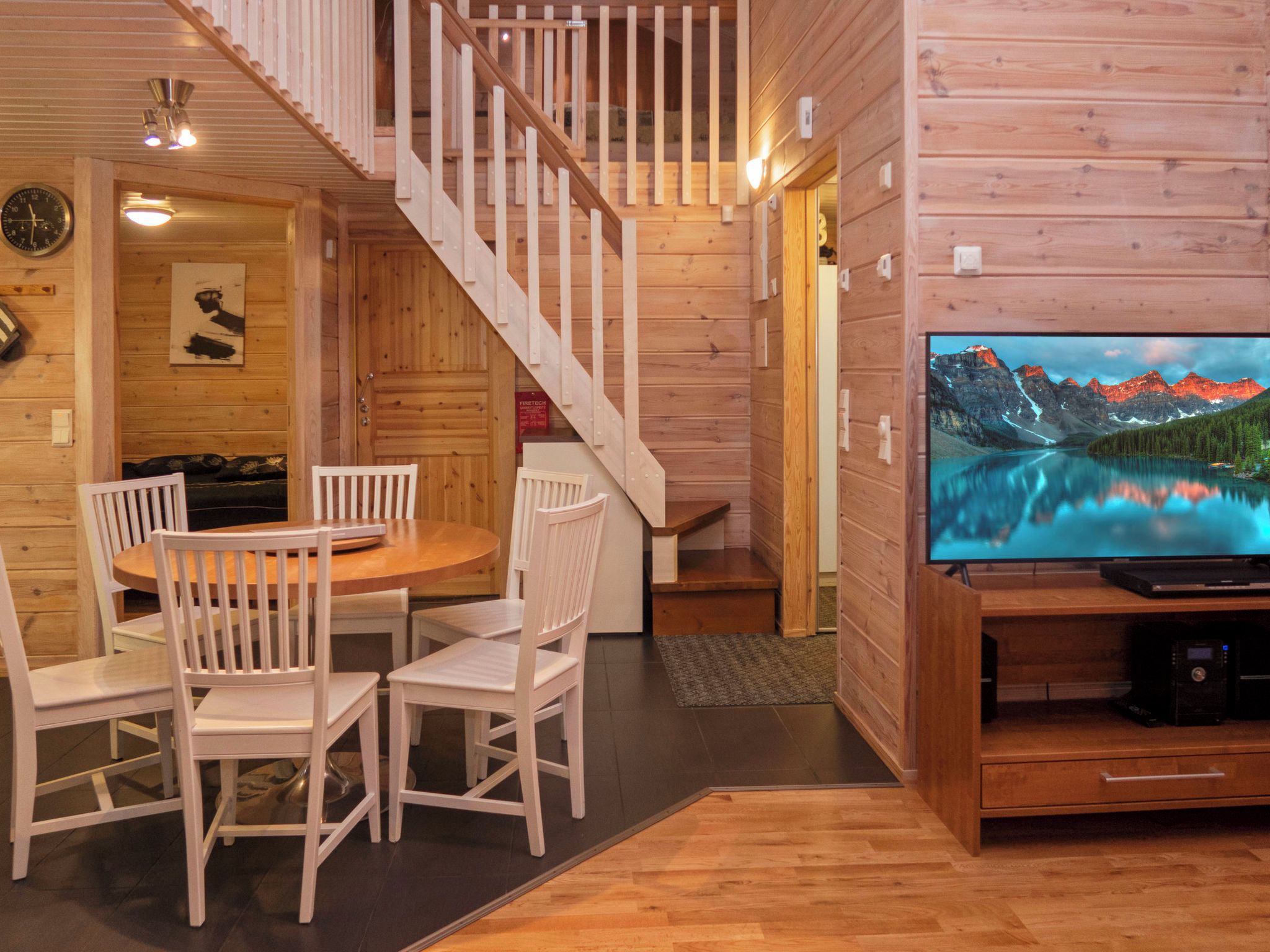 A wooden interior with a dining area, a staircase, and a television displaying a mountain landscape.