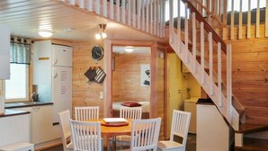 A cozy cabin with a wooden interior, a dining area with white chairs, a kitchen with a refrigerator, and a wooden staircase.