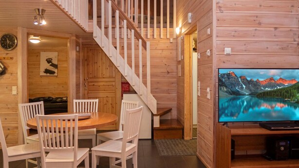 A wooden cabin interior with a dining area, a staircase, and a television displaying a mountain landscape.