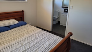 Ein Schlafzimmer mit einem hölzernen Bett, einer gemusterten Bettdecke, einem Fenster mit einem Metallgitter und Blick auf ein Badezimmer durch eine geöffnete Tür.