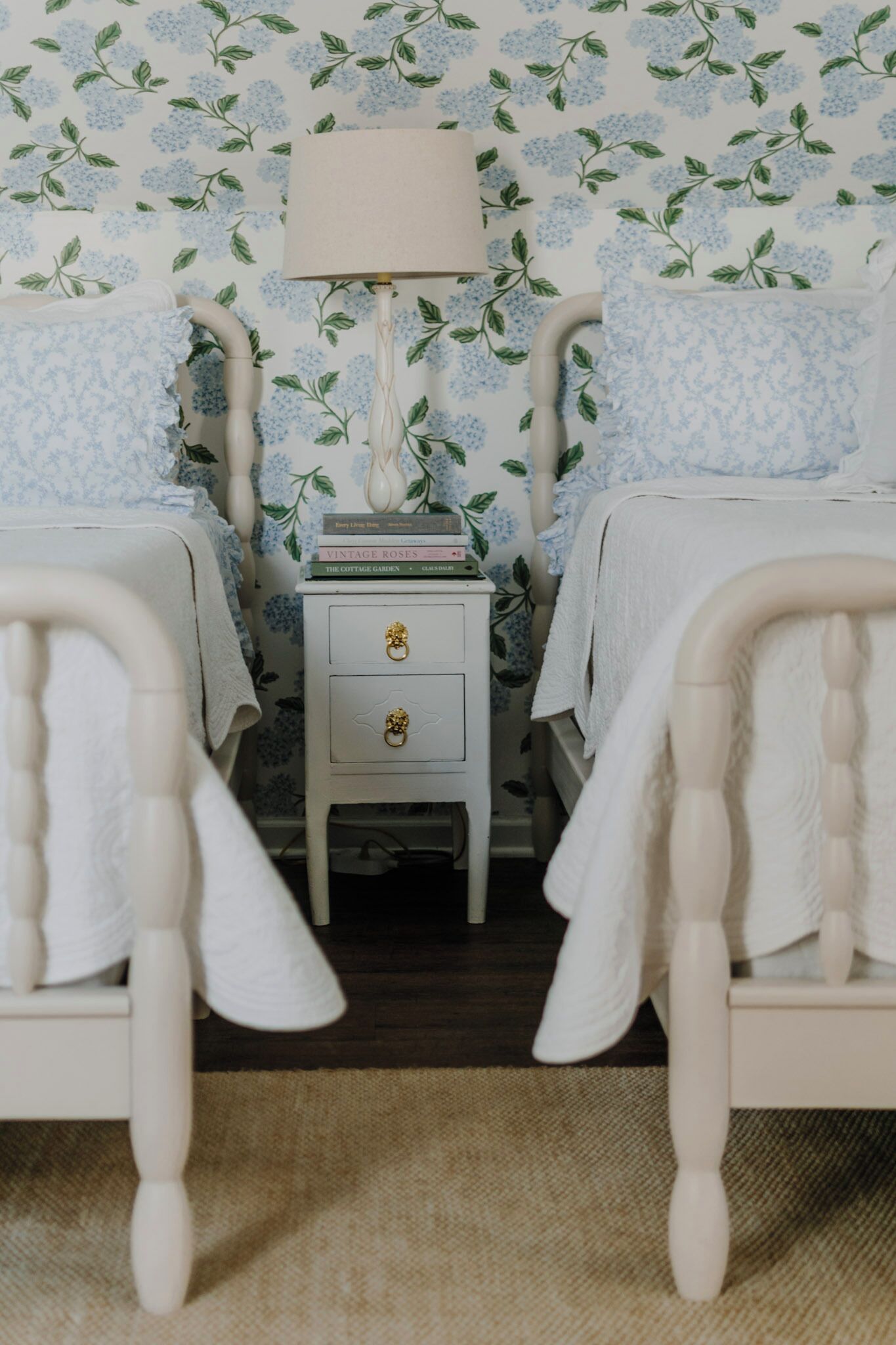 A bedroom with two white beds, a nightstand with a lamp, and a floral wallpaper pattern.