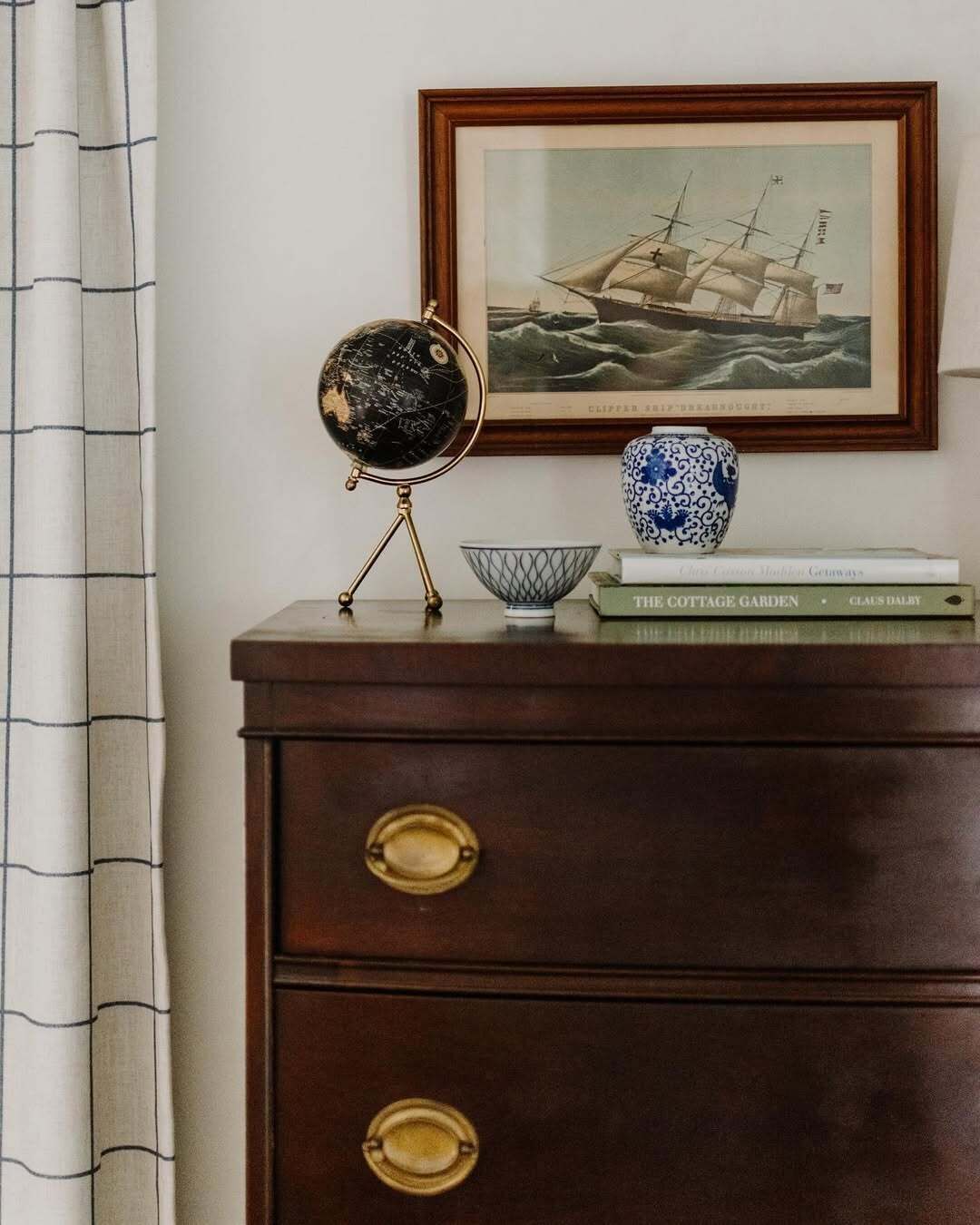 Une commode en bois avec des poignées en laiton, un globe terrestre posé sur un socle, un tableau maritime encadré, un vase bleu et blanc, et un livre intitulé « Le jardin de campagne » de Claus Dalby.